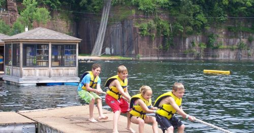 Four children in life jackets pull a rope from a dock over a lake, with a cabin and trees in the background.