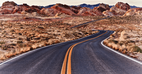 Winding road through a desert landscape with colorful rock formations in the background.