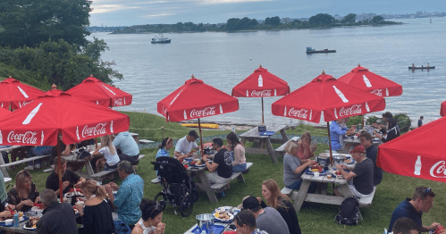 Outdoor dining scene by the water, with people enjoying meals under red umbrellas and boats in the background.