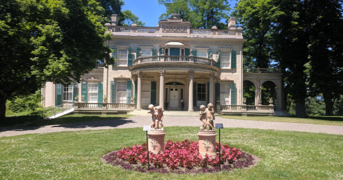 A grand mansion with a balcony, surrounded by trees and two statues in a flowerbed in the foreground.