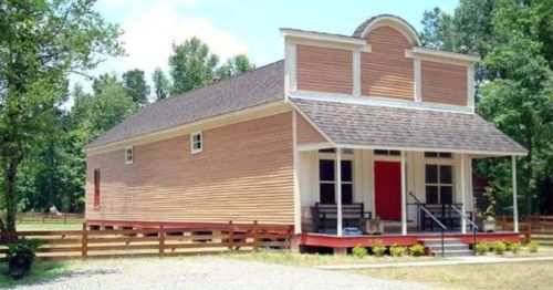 A charming, rustic wooden house with a red door, surrounded by trees and a gravel path.