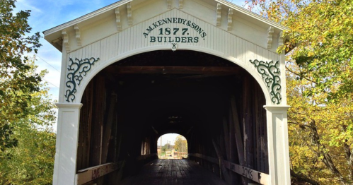 A historic covered bridge with "Kennedy & Sons 1877 Builders" inscribed, surrounded by trees.