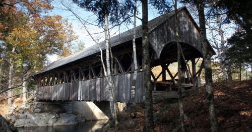 A rustic covered bridge nestled among trees, spanning a small stream with autumn foliage in the background.