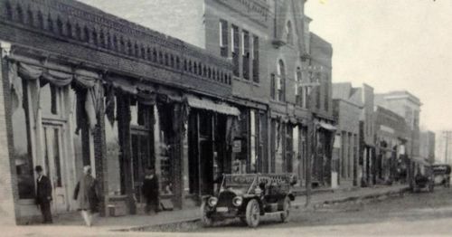 Black and white photo of a historic street with vintage cars and storefronts, showcasing early 20th-century architecture.