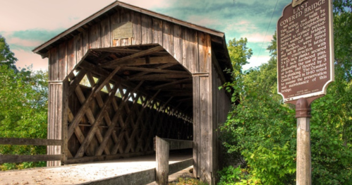 A wooden covered bridge with a sign nearby, surrounded by greenery under a cloudy sky.