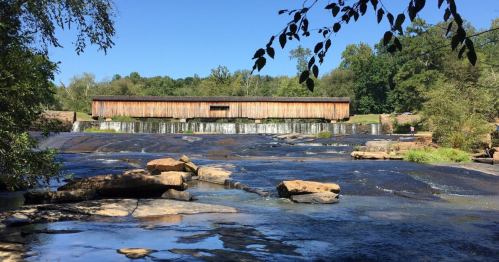 A wooden covered bridge spans a river with gentle waterfalls, surrounded by lush greenery and a clear blue sky.