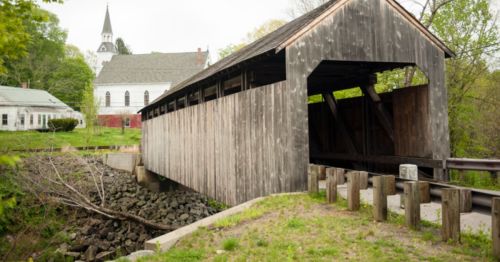A wooden covered bridge spans a small creek, with a church and greenery in the background.
