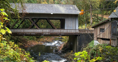 A wooden covered bridge spans a river, surrounded by lush greenery and autumn foliage.