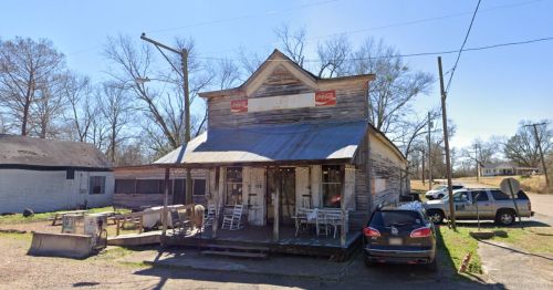 A weathered, rustic storefront with Coca-Cola signs, surrounded by bare trees and parked cars.