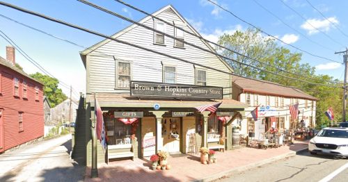 A charming country store with a green exterior, flags, and outdoor seating, surrounded by trees and other buildings.