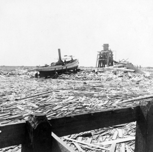 A black and white photo of a boat on a river covered with debris, near a wooden structure in the background.