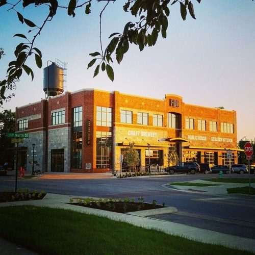 A brick building with large windows, labeled as a craft brewery and public house, surrounded by greenery and parked cars.