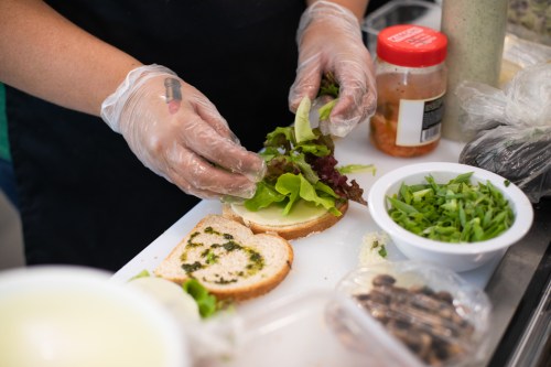 A person in gloves adds fresh lettuce to a sandwich on a cutting board, surrounded by various ingredients.