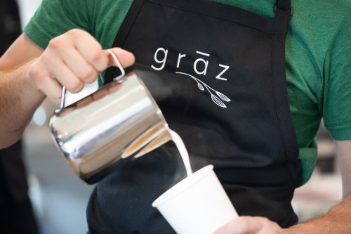 A person in a green shirt pours steamed milk from a metal pitcher into a white cup, wearing an apron with "grāz" logo.