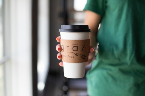 A person holding a coffee cup with a "graz" logo and a brown sleeve, wearing a green shirt.