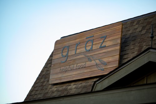 Sign on a building featuring the word "grāz" and the phrase "helpful food" against a clear blue sky.