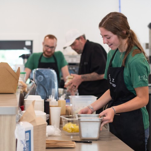 A busy kitchen scene with staff preparing food, wearing aprons and engaged in various tasks at a counter.