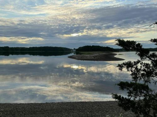 A serene lake scene with a sandy shore, reflecting clouds and trees under a cloudy sky.