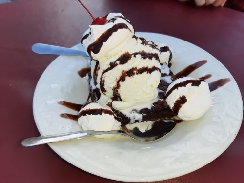 A dessert plate featuring a brownie topped with scoops of vanilla ice cream, chocolate sauce, and a cherry.