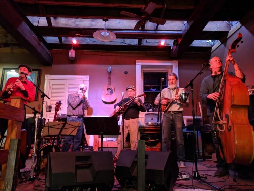 A five-member band performs on stage with various instruments in a cozy venue, featuring wooden beams and warm lighting.