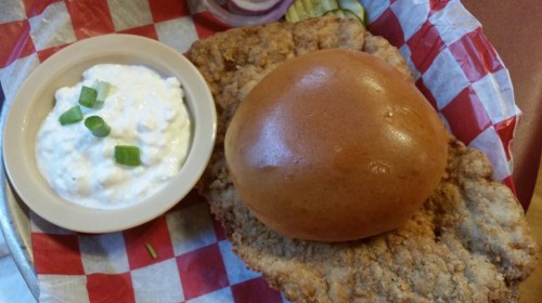 A crispy fried chicken sandwich on a bun, served with a side of creamy dip and garnished with green onions.
