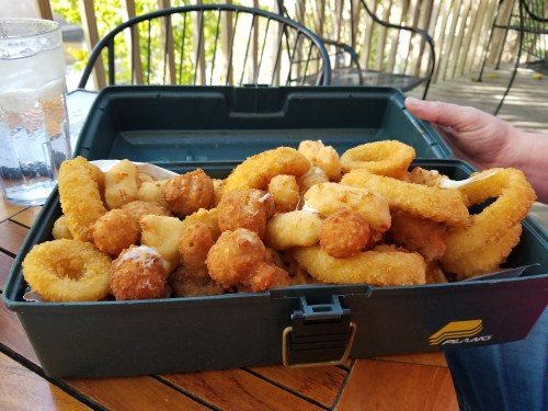 A takeout container filled with various fried seafood, including onion rings and breaded shrimp, on a wooden table.