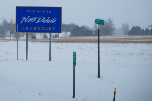 A snowy landscape featuring a "Welcome to North Dakota" sign and road markers indicating distance.