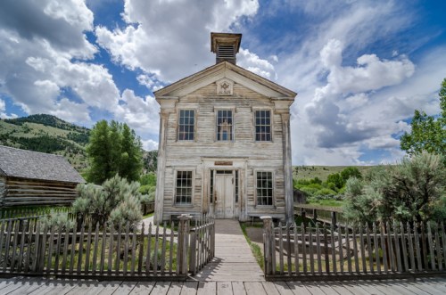 A weathered wooden schoolhouse with a bell tower, surrounded by greenery and under a cloudy sky.
