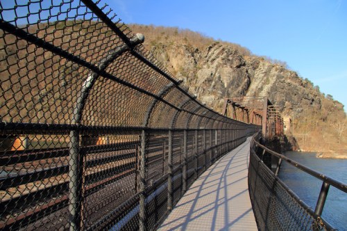 A winding pedestrian bridge with a chain-link fence, surrounded by rocky cliffs and a river under a clear blue sky.