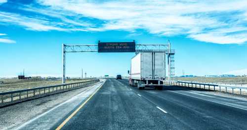 A highway scene with a truck driving under a warning sign for strong wind gusts. Clear blue sky in the background.