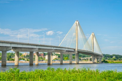 A modern cable-stayed bridge spans a river, with green grass in the foreground and a clear blue sky above.