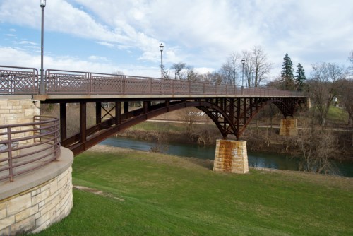 A metal arch bridge spans a river, supported by stone pillars, with grassy banks and trees in the background.