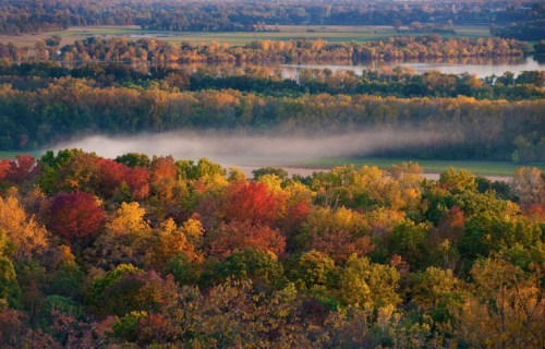 A scenic view of a colorful autumn landscape with trees in vibrant hues and a misty river in the background.
