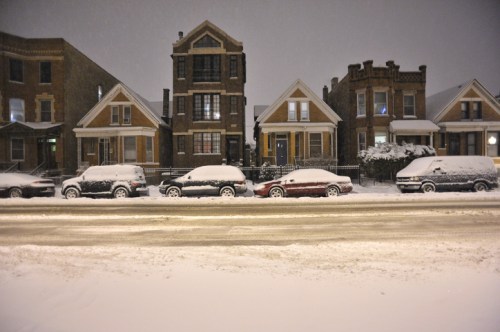Snow-covered cars line a quiet street, with charming houses in the background under a night sky.