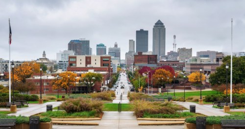 A city skyline with tall buildings, colorful autumn trees, and a wide street under a cloudy sky.