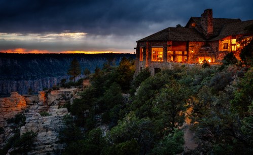 A rustic stone house on a cliff at sunset, surrounded by trees and dramatic clouds over a canyon landscape.