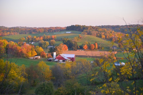 A scenic view of a farm surrounded by colorful autumn trees and rolling hills at sunset.