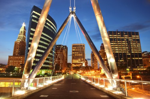 A modern bridge with illuminated supports, leading to a city skyline at dusk, featuring tall buildings and a historic tower.