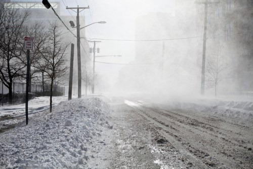 A snowy street with deep snowbanks and blowing snow, creating a foggy, wintry atmosphere.
