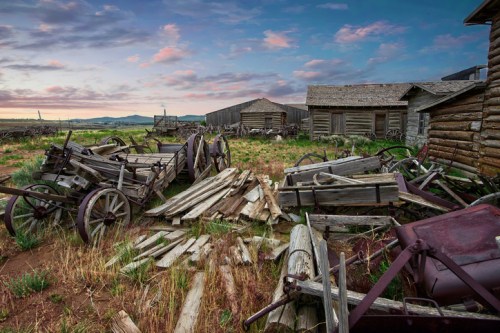 Abandoned wooden structures and old wagons scattered in a grassy field under a colorful sky at sunset.