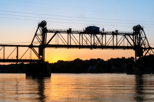 Silhouette of a bridge at sunset, reflecting on calm water with trees in the background.