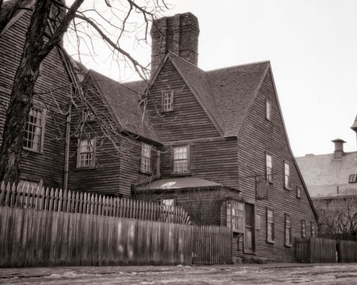 Historic wooden house with a chimney, surrounded by a picket fence and bare trees, in a black-and-white setting.