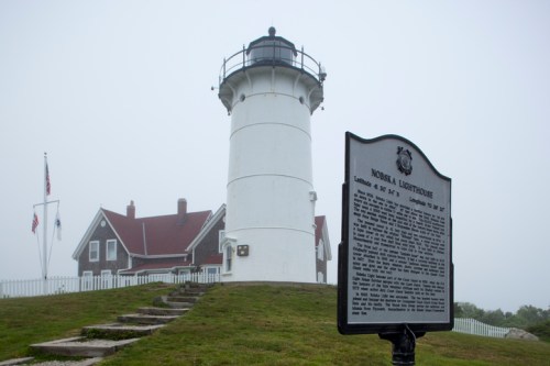 A white lighthouse with a red-roofed building nearby, shrouded in fog, alongside a historical marker.