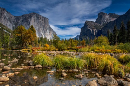 A serene landscape featuring a river, rocky shore, and towering cliffs, surrounded by colorful autumn foliage and blue skies.