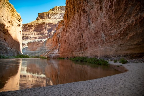 A serene river flows between towering red rock cliffs under a clear blue sky.