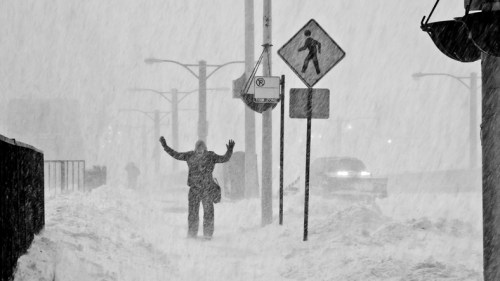 A person stands with arms raised in a heavy snowstorm, surrounded by snow and obscured street signs.