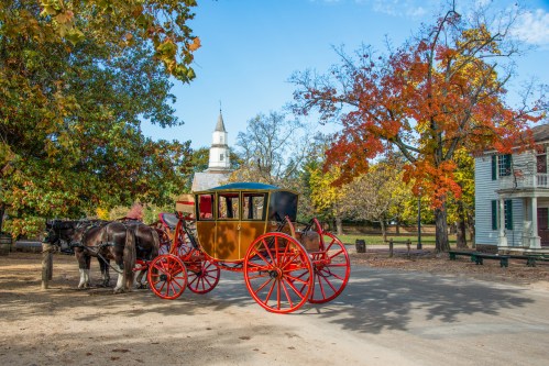 A colorful horse-drawn carriage near trees with autumn leaves and a church steeple in the background.