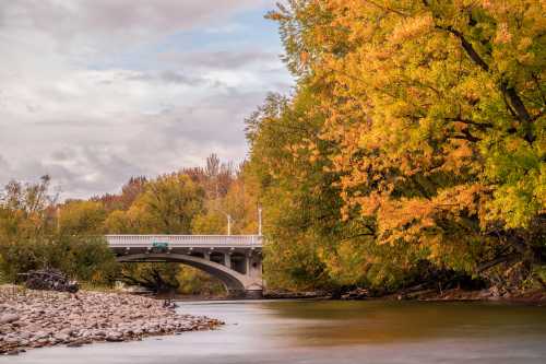 A serene river scene with autumn foliage and a bridge in the background under a cloudy sky.