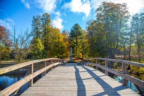 A wooden bridge leads to a monument surrounded by colorful autumn trees under a bright blue sky.
