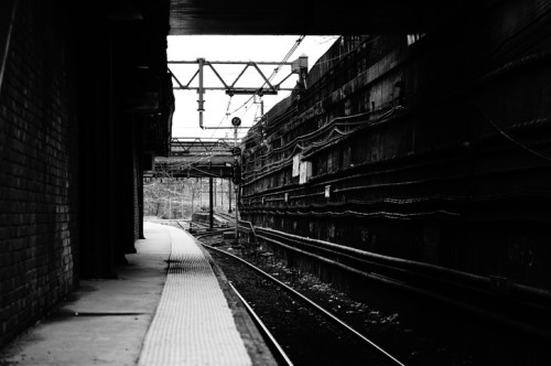 A black and white image of a train platform with tracks and industrial structures in the background.